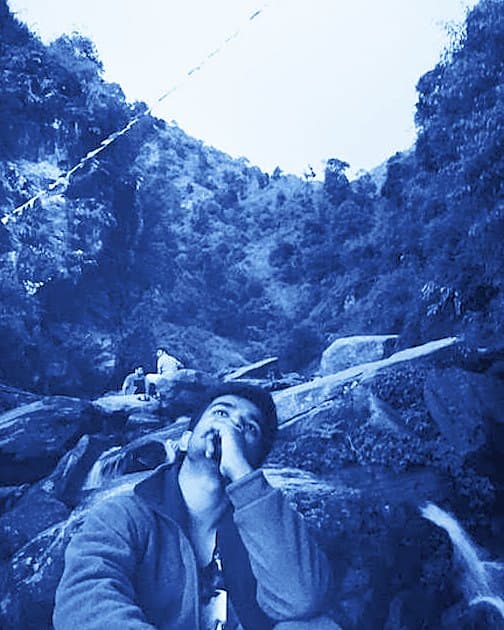 Bidhan Gupta sitting on a rock by a mountain stream, looking up at prayer flags strung across the valley